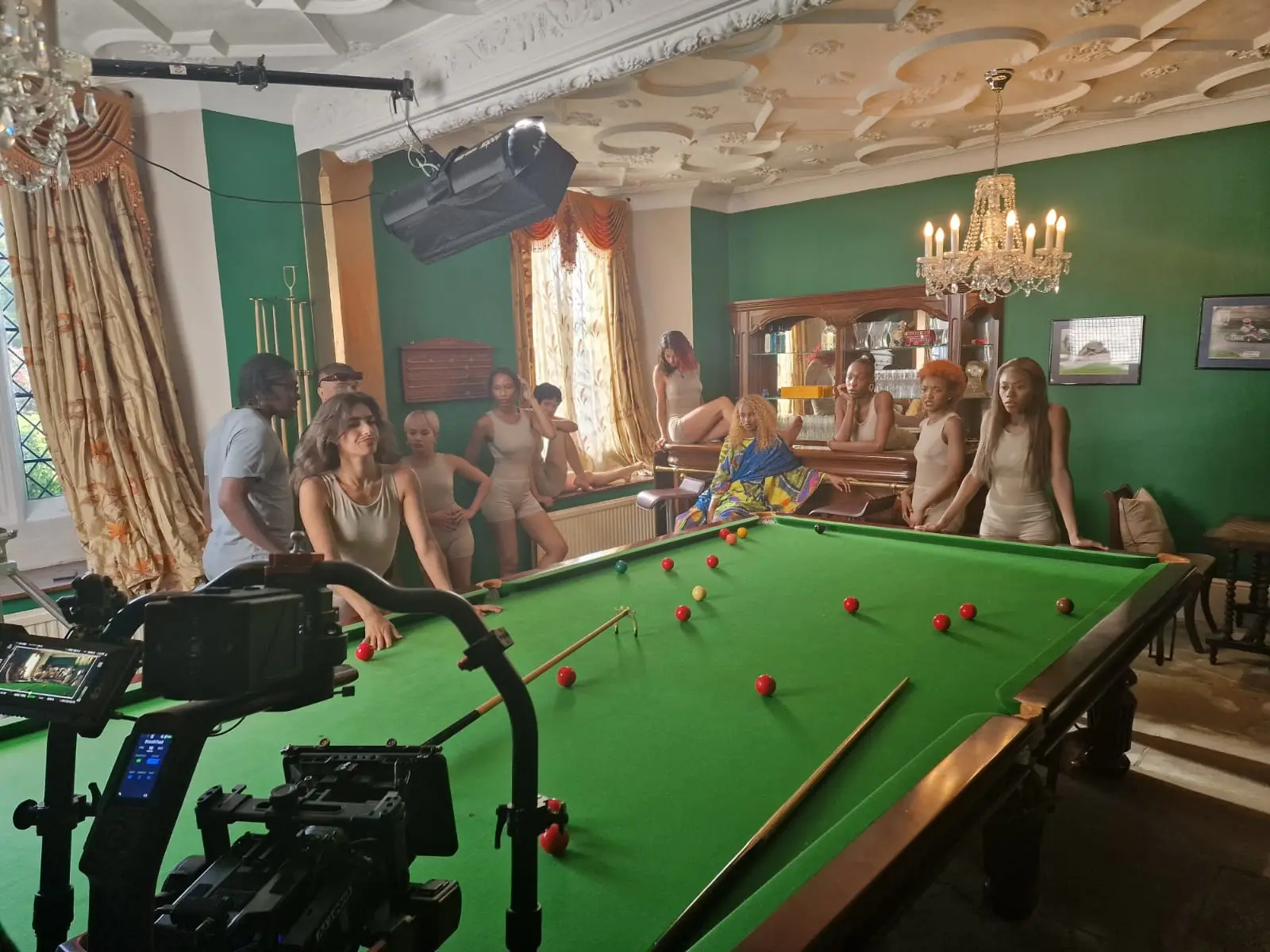 A film crew and camera rig in the billiard room, with cast around the snooker table under a chandelier