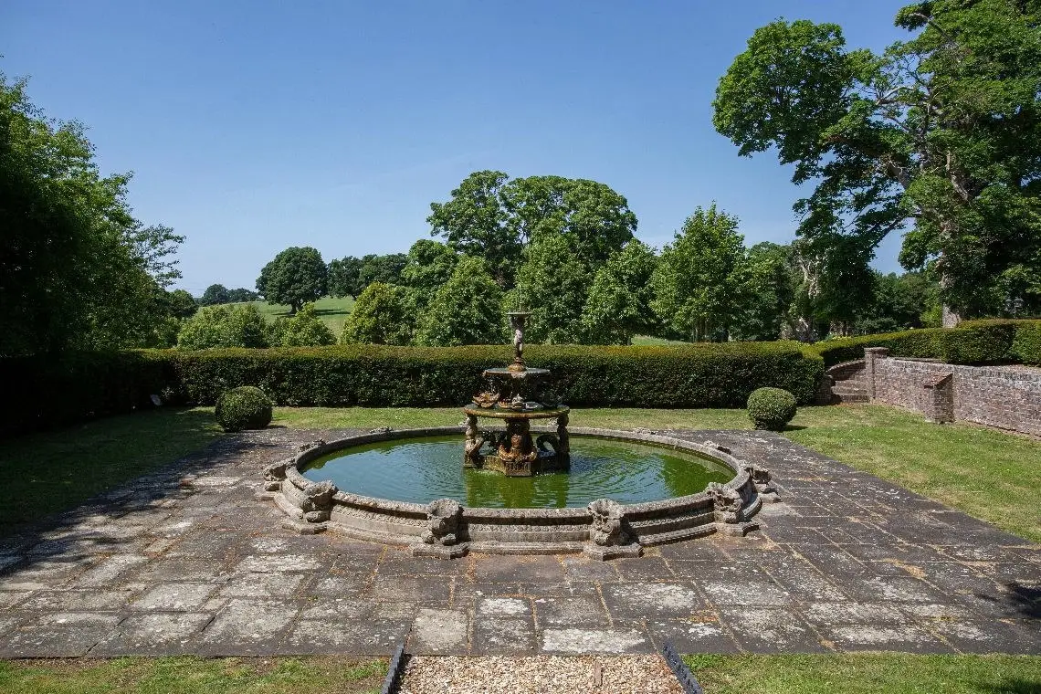 A circular fountain pool in a paved court enclosed by hedges