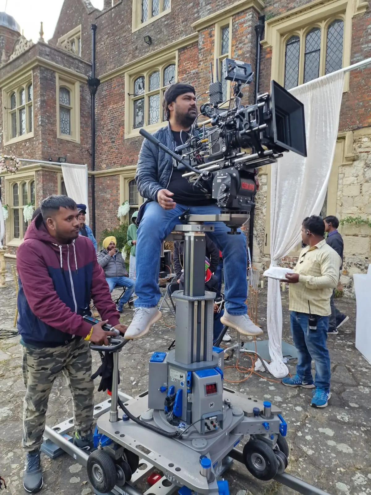 A camera operator on a dolly in front of the brick garden front of the house